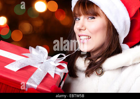 Junge fröhliche Frau Weihnachtsmann Mütze und Handschuhe hält Weihnachtsgeschenk umhüllt von rotem Papier, auf Hintergrund unscharf Stockfoto