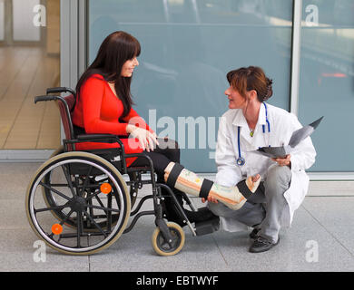 junge Frau mit Bein in Gips in einem Rollstuhl sitzen und im Gespräch mit einem Arzt Stockfoto