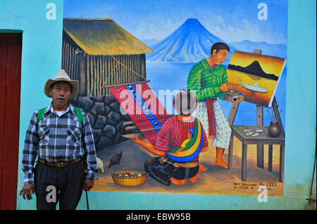 Mann steht vor einem Gemälde an einer Hauswand zeigt einen Mann malen ein Bild und eine Frau, die Weberei eines Teppich, Guatemala, Atitlan Sees, Santa Cruz la Laguna Stockfoto