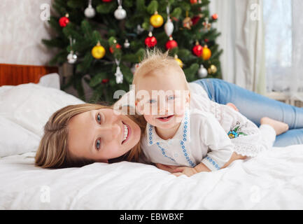 Porträt der glückliche Mutter und Baby Boy auf dem Bett zu Hause mit geschmückter Weihnachtsbaum im Hintergrund Stockfoto