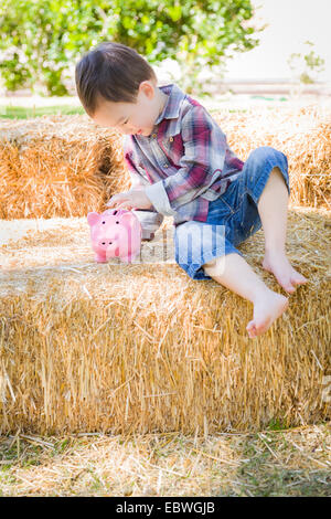 Süße junge gemischte Rennen Junge sitzt auf Heuballen rosa Sparschwein Münzen Inbetriebnahme. Stockfoto