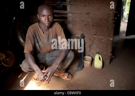 junger Mann hocken in den Eingang von seinem bescheidenen Lehmhaus mit einer Machete neben ihm, Burundi, Karuzi, Buhiga Stockfoto
