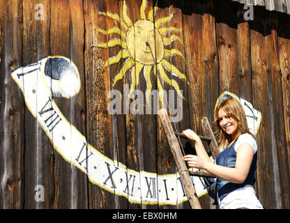 lächelnde langhaarige blonde junge Frau auf einer Leiter stehend lehnte sich gegen eine Palisade Graben mit einer Sonnenuhr gemalt Stockfoto