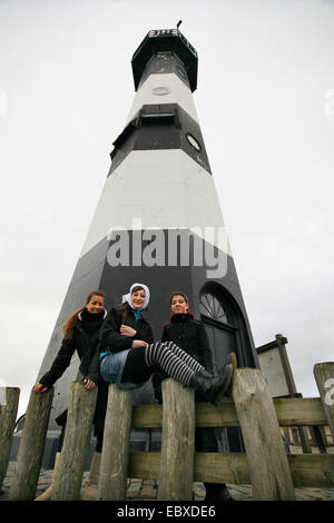 drei junge Frauen sitzen auf Holzzaun vor einem Leuchtturm, Niederlande, Zeeland, Breskens, Sluis Stockfoto