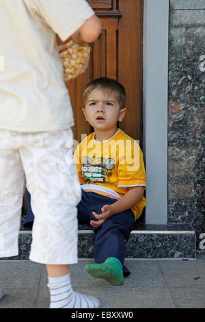 kleiner Junge sitzt auf der Treppe vor einer Tür, ein weiteres Kind fragend anschauen Stockfoto