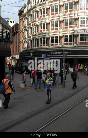 Sonnigen Porträt Menschen überqueren Tib Street Market Street, mit männlichen Verkäufer verkaufen großes Problem im Norden Magazin, Manchester Stockfoto