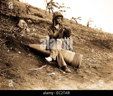 Über eine nicht explodierte 16-Zoll naval Shell für eine Ruhestätte, marine Pfc. Raymond Hubert, schüttelt eine dreitägige Ansammlung von Sand aus seinem boondocker. Saipan, Juli 4, 1944. Stockfoto