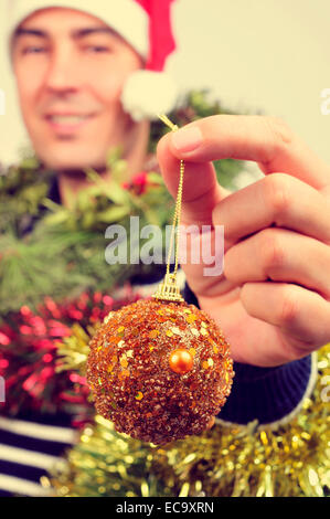 ein junger Mann einen Santa Hut bedeckt mit Christbaumschmuck und eine Kugel in seiner Hand hält Stockfoto