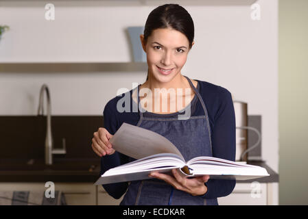 Hübsche junge Frau in eine Schürze, ein Rezept in ein großes gebundenes Buch nachschlagen, während sie in ihrer Küche bereitet sich c steht lächelnd Stockfoto