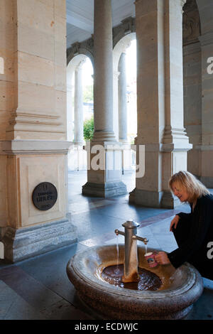 Blonde Frau füllt einen typische Keramik-Becher mit Mineral-Thermalwasser in einem der fünf Quellen am Mühlenkolonnade, Karlovy Vary Stockfoto