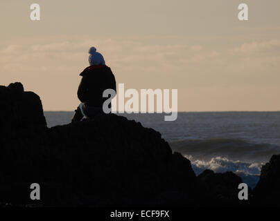 Teenager-Mädchen tragen eine Pudelmütze saß auf Felsen, Blick auf das Meer, Bude, Cornwall, UK Stockfoto