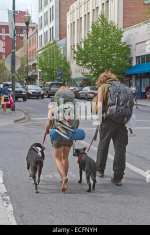 Ein junger Mann und eine Frau auf der Straße tragen Rucksäcke und zwei Hunden an der Leine spazieren überqueren einer Stadtstraße Asheville, NC Stockfoto