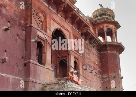 Zwei Babas in traditionellen orangefarbenen Gewändern sitzen auf den Stufen des einen Hindu-Tempel in Varanasi.  Tauben fliegen overhead in der Nachmittagssonne Stockfoto