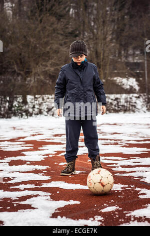 kleiner Junge mit schweren Ausdruck stand vor Ball auf einem roten Platz Stockfoto