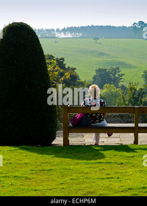 Ältere Frau in einem Garten sitzen auf einer Bank entspannen und genießen der Aussicht auf ländliche Landschaft neben Formschnitt Baum Stockfoto