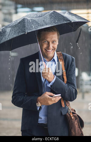 Porträt von lächelnden Geschäftsmann mit Smartphone in der Stadt während der Regenzeit Stockfoto