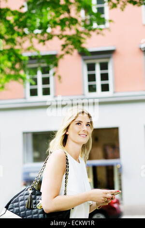 Junge Frau wegsehen gedrückter Handy gegen Gebäude Stadt Stockfoto