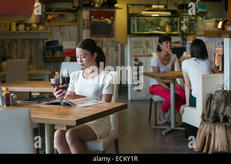 Frau sitzt an einem Tisch in einem Café, Blick auf ihr Handy, zwei Frauen an einem Tisch im Hintergrund. Stockfoto
