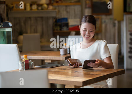 Frau sitzt an einem Tisch in einem Café, hält die Rechnung und ihre Geldbörse, lächelnd. Stockfoto