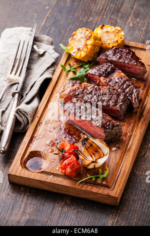 In Scheiben geschnitten mittlere seltene Gegrilltes Beefsteak Ribeye mit Mais und Kirschtomaten auf Schneidebrett auf hölzernen Hintergrund Stockfoto