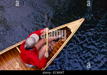 Eine junge Frau in einem leuchtend roten Jacke Paddel Vintage Holz Kanu in einem stream Stockfoto
