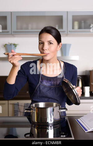 Junge Hausfrau, die ihre Küche wie sie das Abendessen in ihrer Küche Probenahme direkt aus dem Topf auf dem Herd mit einem w bereitet Stockfoto