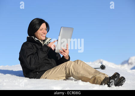 Glückliche Wanderer Frau Surfen eine Tablette auf dem Schnee mit dem Himmel im Hintergrund sitzen Stockfoto