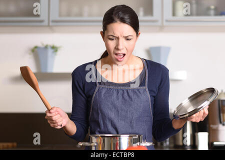 Junge Hausfrau mit einem Unglück in der Küche in Schock und das Entsetzen zu reagieren, wie sie den Deckel auf den Topf auf dem Herd hebt Stockfoto