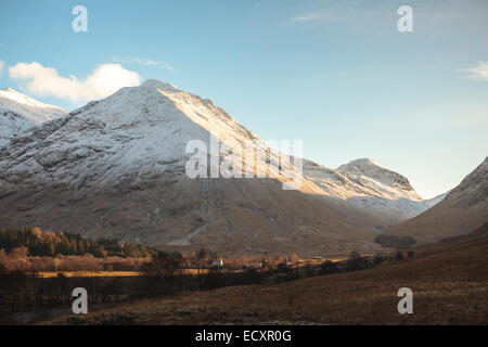 Nachmittagssonne auf den schneebedeckten vulkanischen Gipfeln von Glen Coe, Schottland, in den schottischen Highlands. Ein Bauernhaus wird durch das Sonnenlicht beleuchtet. Stockfoto