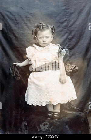 Ca. 1900 Schwarz-Weiß-Fotografie. Studioportrait eines unglücklich aussehenden zweijährigen Jungen, der in seinem Taufkleid mit Stoffhintergrund steht. Stockfoto