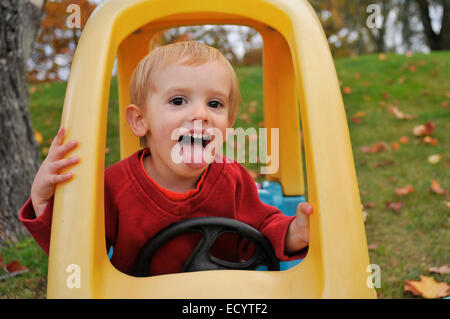 Ein zwei-Jahr-alte junge macht ein lustiges Gesicht während des Spielens in seinem Spielzeugauto an einem Herbsttag. Stockfoto