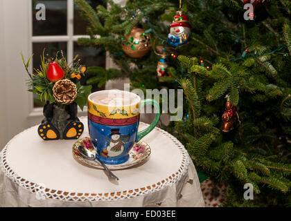 Weihnachten-Becher mit Heißgetränk auf kleinen Tisch durch einen Weihnachtsbaum.  Es ist ein schwarzer Keramik Bär, eine Weihnachten Blume Stockfoto