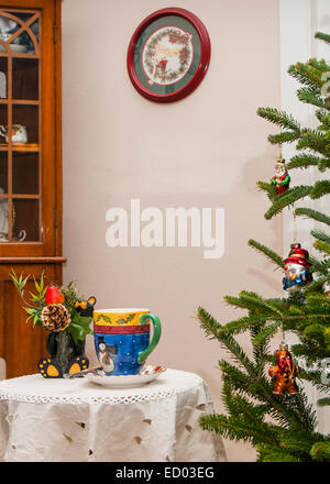 Weihnachten-Becher mit Heißgetränk auf kleinen Tisch durch einen Weihnachtsbaum.  Es ist ein schwarzer Keramik Bär, die eine Blume. Stockfoto