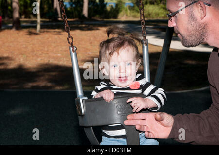 Ein Kleinkind Mädchen in einem Park in einer Babyschaukel.  Ihr Vater drängt sie von vorne.  Sie lächelt. Stockfoto