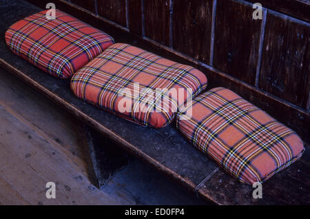 Drei verblasste rote Tartan Knie- oder Sitzkissen oder Kissen liegen auf dunklen Holzsitz oder Pew mit Holzboden Stockfoto