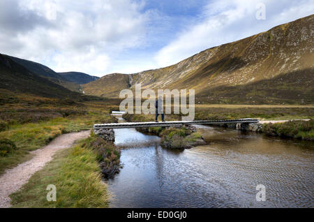 Eine weibliche Walker überquert einen Holzsteg der Allt ein Dubh Loch mit den Hängen des weißen Mounth im Hintergrund Stockfoto