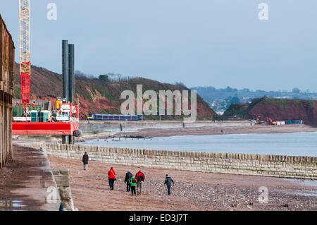 Menschen zu Fuß entlang des Strandes in Dawlish mit dem Riesen reparieren Rig stand neben der beschädigten Ufermauer im Hintergrund Stockfoto