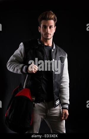 Wunderschönen Mann in trendigen Mode Outfit tragen University Jacke mit Sporttasche hautnah. Aufgenommen im Studio auf schwarzem Hintergrund. Stockfoto