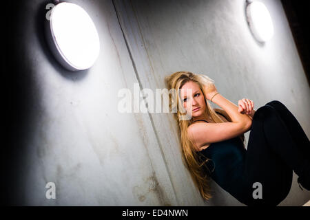 Eine junge Frau (18 Jahre altes Mädchen) mit langen blonden Haaren, solo allein, suchen nachdenklich nachdenklich einsam sitzen mit dem Rücken gegen eine Betonwand in ein Outdoor-urbaner Backstreet Citylage, UK Stockfoto