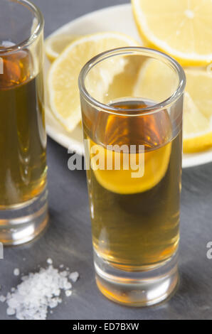 Tequila in einem Schnapsglas, grobes Salz auf Schiefer und Zitrone Scheibe auf einer Platte im Hintergrund Stockfoto