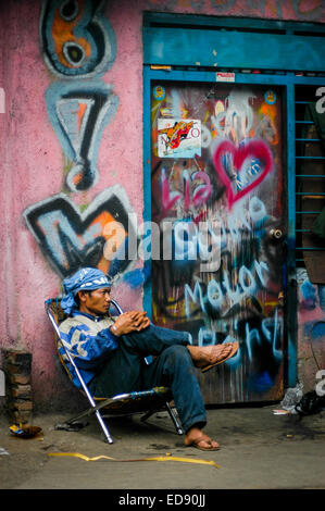 Ein Mann mit Bandana sitzt vor einem Haus am Straßenrand, das mit Graffiti dekoriert ist, in einem dichten Viertel in Jakarta, Indonesien. Stockfoto