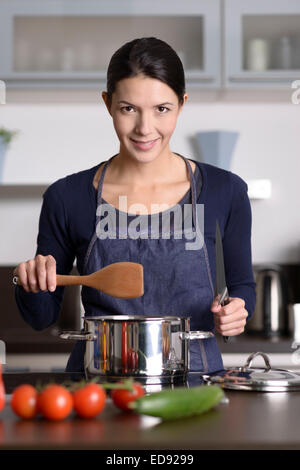 Glückliche freundliche junge Hausfrau kochen Abendessen steht auf dem Kochfeld einen Holzlöffel über einem Topf halten, wie sie die Fre bereitet Stockfoto