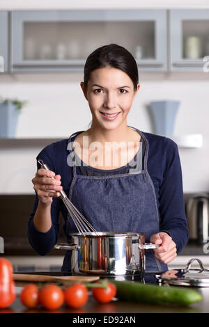 Glückliche freundliche junge Hausfrau kochen Abendessen steht auf dem Kochfeld, einen Schneebesen über einem Topf halten, wie sie das Essen bereitet Stockfoto