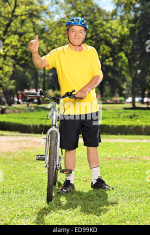 In voller Länge Portrait von einem nahen im Alter Mann im Daumen aufgeben, neben seinem Fahrrad in einem Park Sportbekleidung Stockfoto