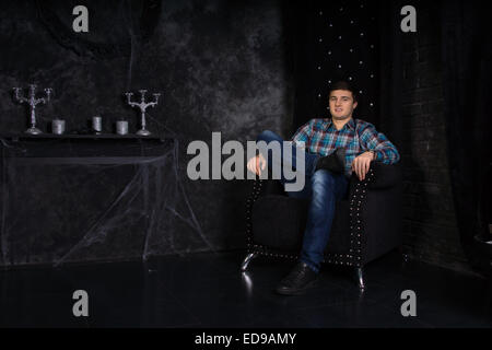 Lächelnde junge Mann, sitzend mit Beine gekreuzt in Plüsch schwarz hoher Stuhl zurück in unheimliche Spukhaus Einstellung mit Spinnweben Stockfoto