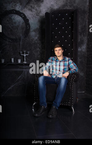 Mann trägt Freizeitkleidung zurücklehnen in High Chair in schaurigen Halloween Haunted Haus Einstellung mit Kandelaber und Spinnweben Stockfoto