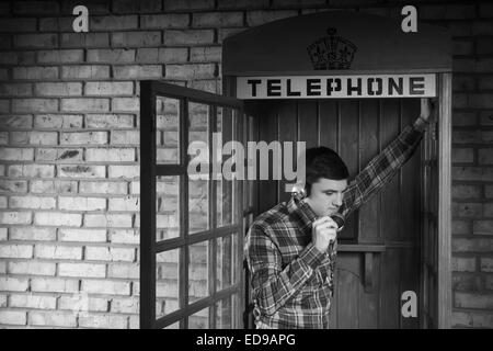 Junger Mann ruft jemand an die Telefonzelle mit Ziegel Wand Hintergrund. In Monochrom erfasst. Stockfoto