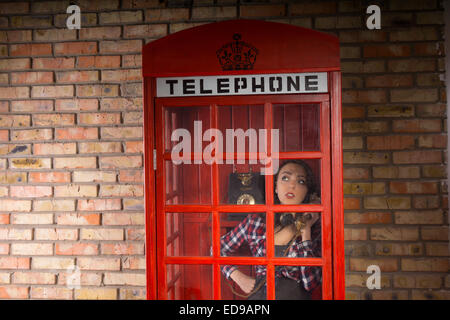 Junge Frau im Gespräch jemand ernsthaft in einer geschlossenen roten Telefonzelle mit einem Brick Wall Hintergrund. Stockfoto