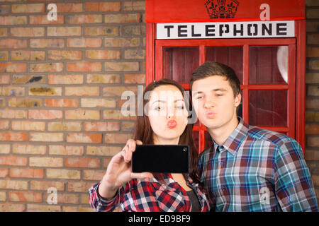Junges Paar posieren vor eine ikonische rote britische Telefonzelle nehmen eine Selfie von sich selbst auf ihrem Handy beim ziehen Stockfoto