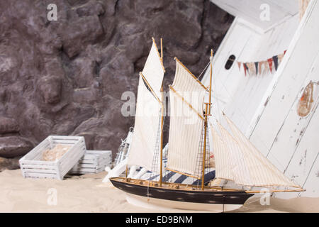 Spielzeug-Yacht und strandete Holzschiff in marine oder nautischen Stillleben arrangiert ein Sandstrand am Fuße einer Felswand mit Stockfoto